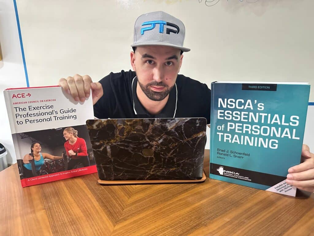 ACE vs NSCA - Tyler holds up ACE and NSCA textbooks on a table with laptop between them in front of a whiteboard