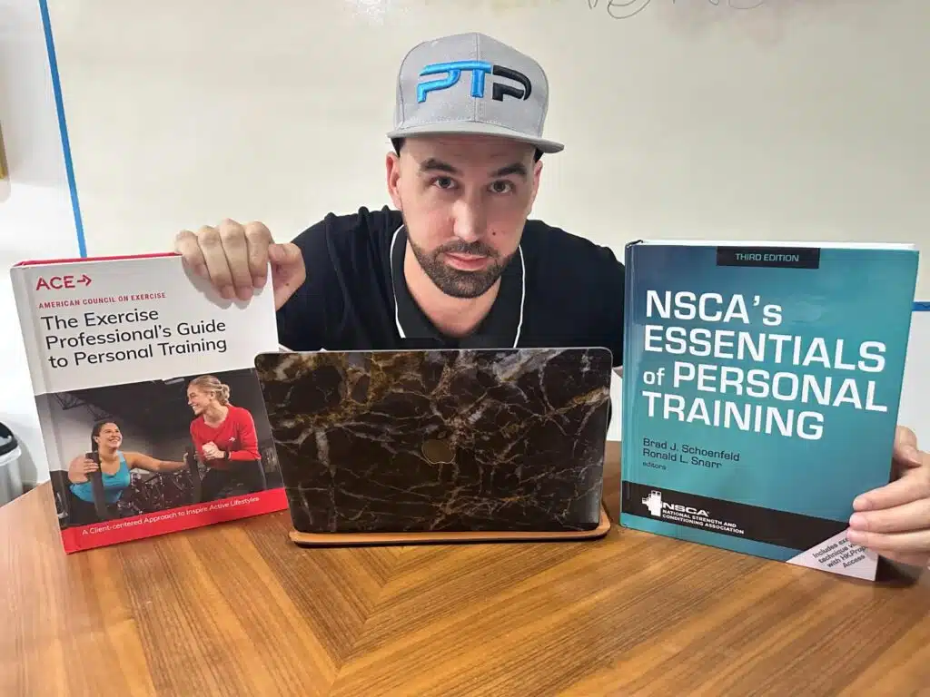 ACE vs NSCA - Tyler holds up ACE and NSCA textbooks on a table with laptop between them in front of a whiteboard