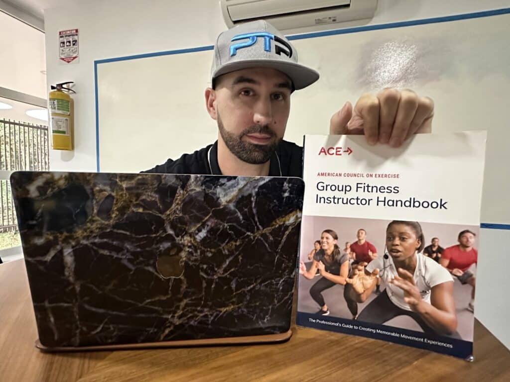 ACE Group Fitness Instructor review - Tyler read holds up ace textbook on a table next to a laptop 