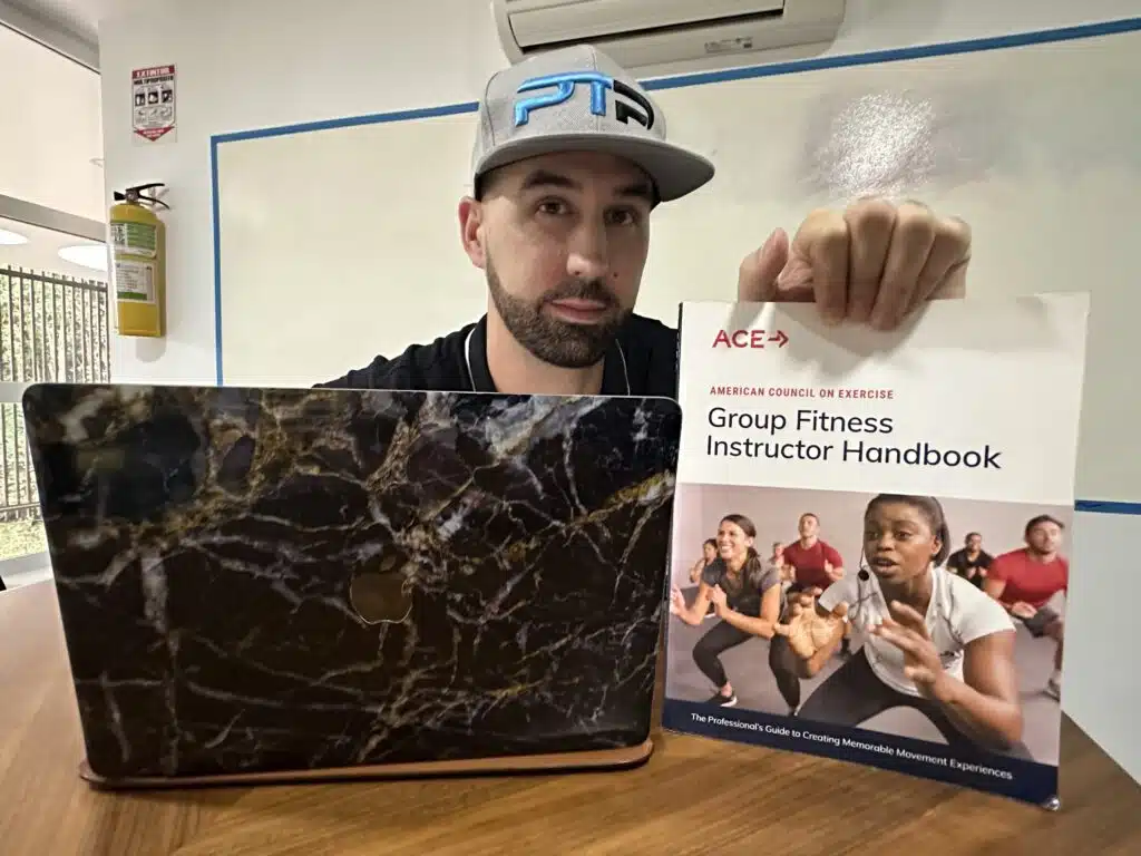 The Ultimate ACE Group Fitness Instructor Review 1 ACE Group Fitness Instructor review - Tyler read holds up ace textbook on a table next to a laptop