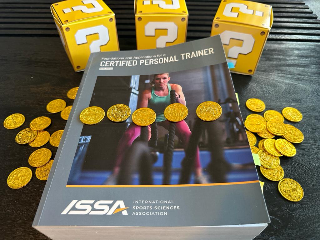 Cheapest personal training certifications. ISSA textbook laid out on table with gold coins and mystery boxes - how much does ISSA CPT cost?