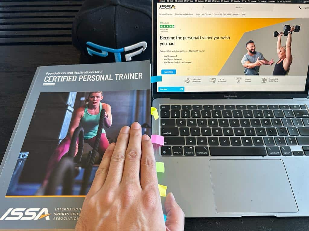 Best online personal training certification - ISSA textbook next to laptop with the issa website on the screen - ptpioneer hat and hand visible