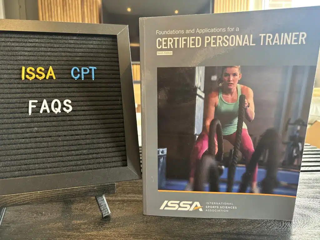 ISSA Training Certification Review 2023 - Is ISSA Worth It? 24 ISSA trainer certification frequently asked questions (FAQs) - ISSA Certified Personal Trainer textbook displayed on table with ISSA FAQs on letterboard